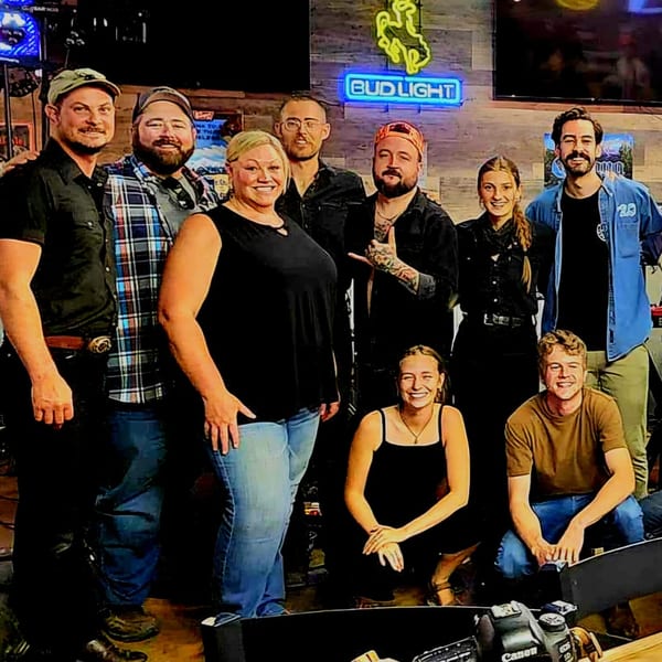 Nine smiling Brewgards Lounge staff members pose together under a Bud Light neon sign.