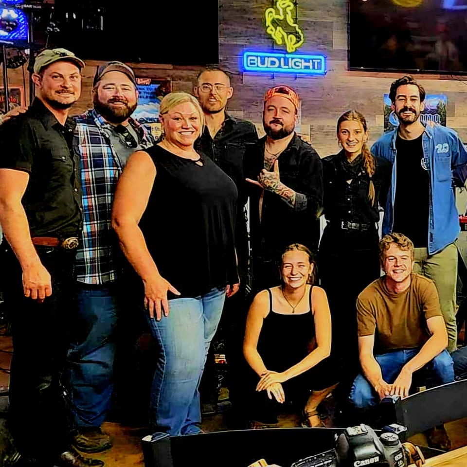 Nine smiling Brewgards Lounge staff members pose together under a Bud Light neon sign.