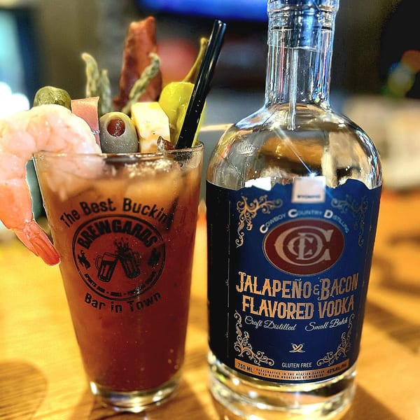 A Brewgards Bloody Mary garnished with shrimp, olives, and cheese, next to a bottle of Jalapeño & Bacon Vodka.