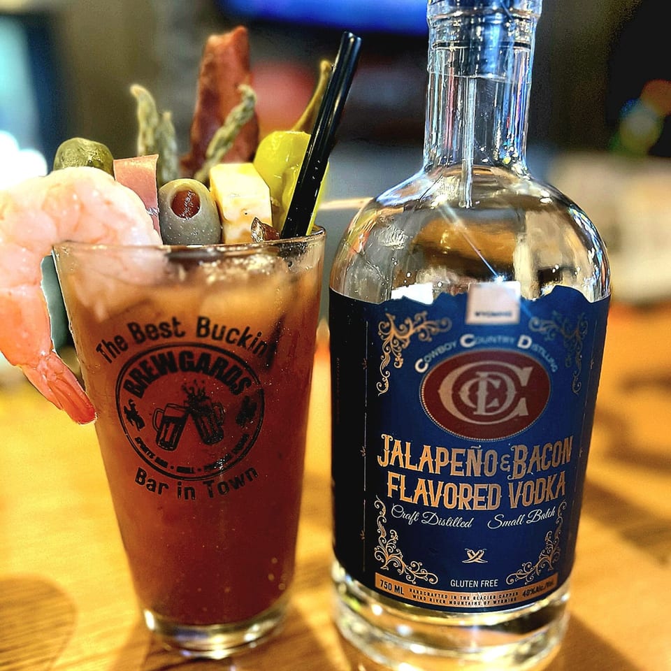 A Brewgards Bloody Mary garnished with shrimp, olives, and cheese, next to a bottle of Jalapeño & Bacon Vodka.