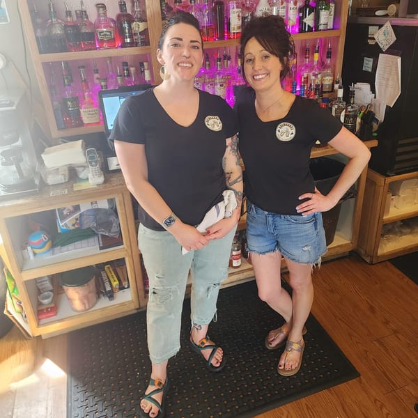 Two smiling Brewgards Lounge bartenders stand in front of a liquor shelf with pink lighting.