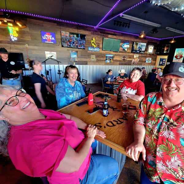 Patrons smiling at a high-top table in Brewgards Lounge, with neon signs and TVs on the wall.
