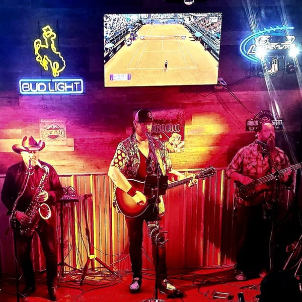 Three musicians performing live on a stage with neon signs and a large TV screen showing tennis.